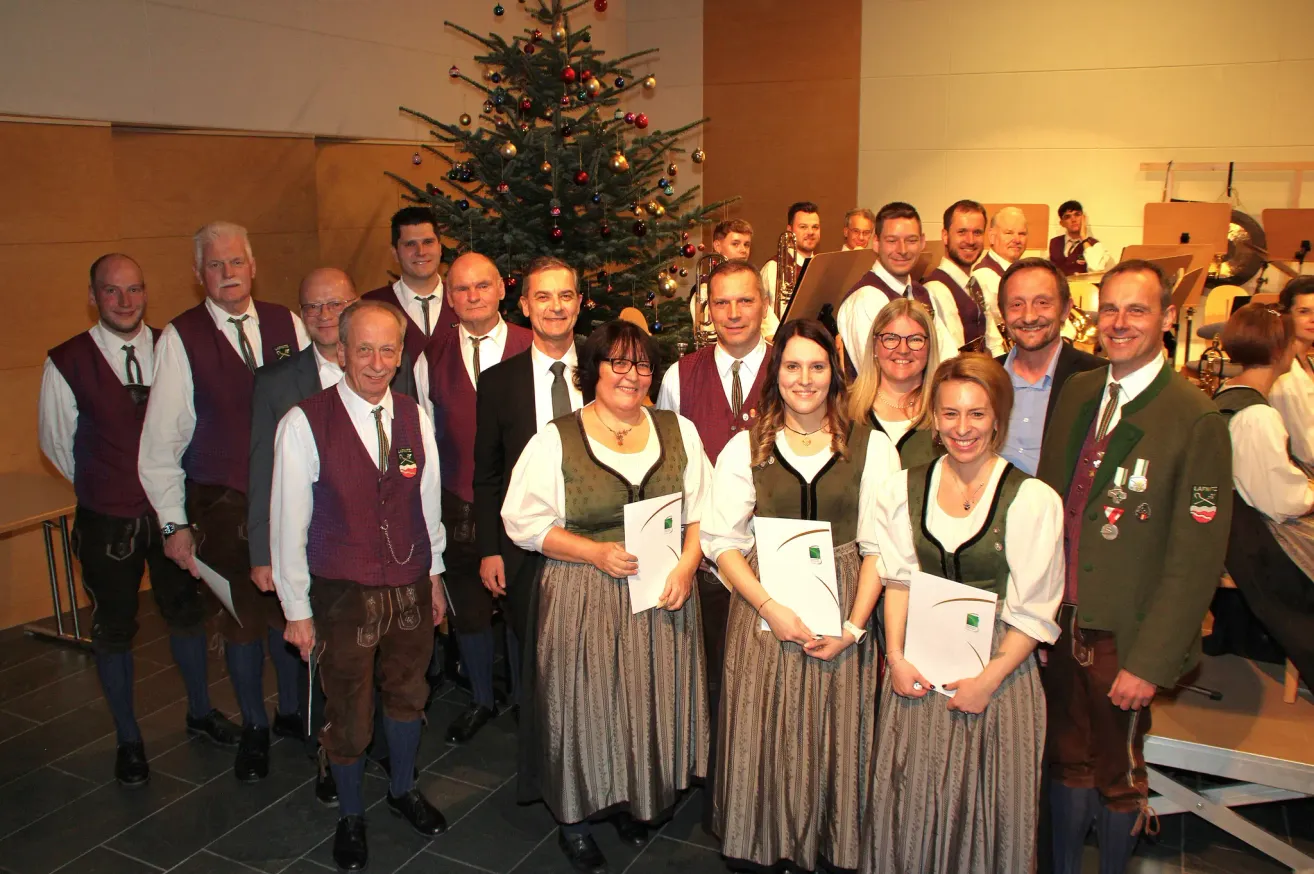 Eine Gruppe von Menschen in traditioneller Kleidung steht lächelnd vor einem geschmückten Weihnachtsbaum, Papiere in der Hand. Dahinter sind Musiker zu sehen, einige spielen Instrumente.