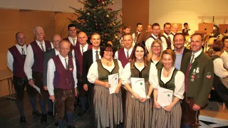 Eine Gruppe von Menschen in traditioneller Kleidung steht lächelnd vor einem geschmückten Weihnachtsbaum, Papiere in der Hand. Dahinter sind Musiker zu sehen, einige spielen Instrumente.