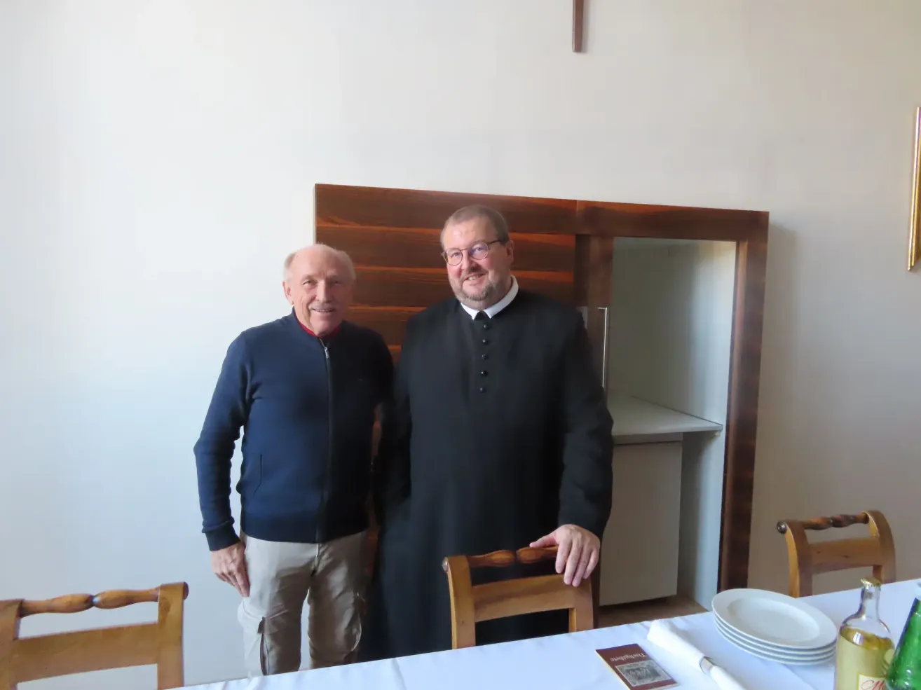 Zwei Männer stehen in einem Raum mit einer Holzwand. Der Mann auf der linken Seite trägt eine blaue Jacke und Khakihosen. Der Mann auf der rechten Seite trägt einen schwarzen Gewand mit einem weißen Kragen. Sie lächeln beide. Hinter ihnen steht ein Tisch mit Tellern und einem Buch.