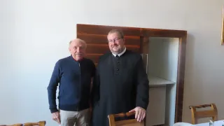 Zwei Männer stehen in einem Raum mit einer Holzwand. Der Mann auf der linken Seite trägt eine blaue Jacke und Khakihosen. Der Mann auf der rechten Seite trägt einen schwarzen Gewand mit einem weißen Kragen. Sie lächeln beide. Hinter ihnen steht ein Tisch mit Tellern und einem Buch.