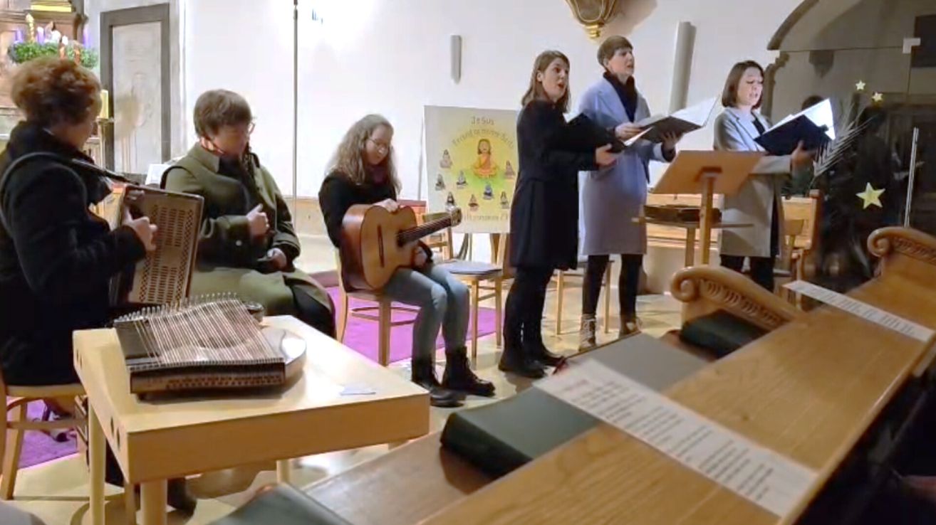 Vier Frauen in einem Raum, eine spielt Gitarre, andere singen und lesen aus einem Buch, mit einem Poster im Hintergrund.