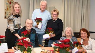 Vier ältere Personen sitzen um einen Tisch herum mit Blumen als Tischdekoration und einem Glas Champagner. Ein Mann hält ein Geschenk und eine Frau trägt eine Brille und eine Perlenkette.