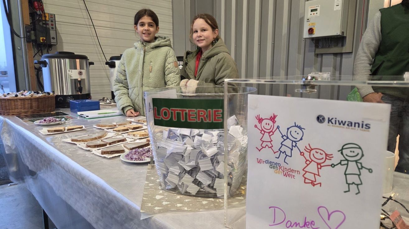 Zwei Mädchen stehen hinter einem Tisch mit Essen und einem Lotterie-Schild. Ein Glas voller Zettel ist mit 'Lotterie' beschriftet.