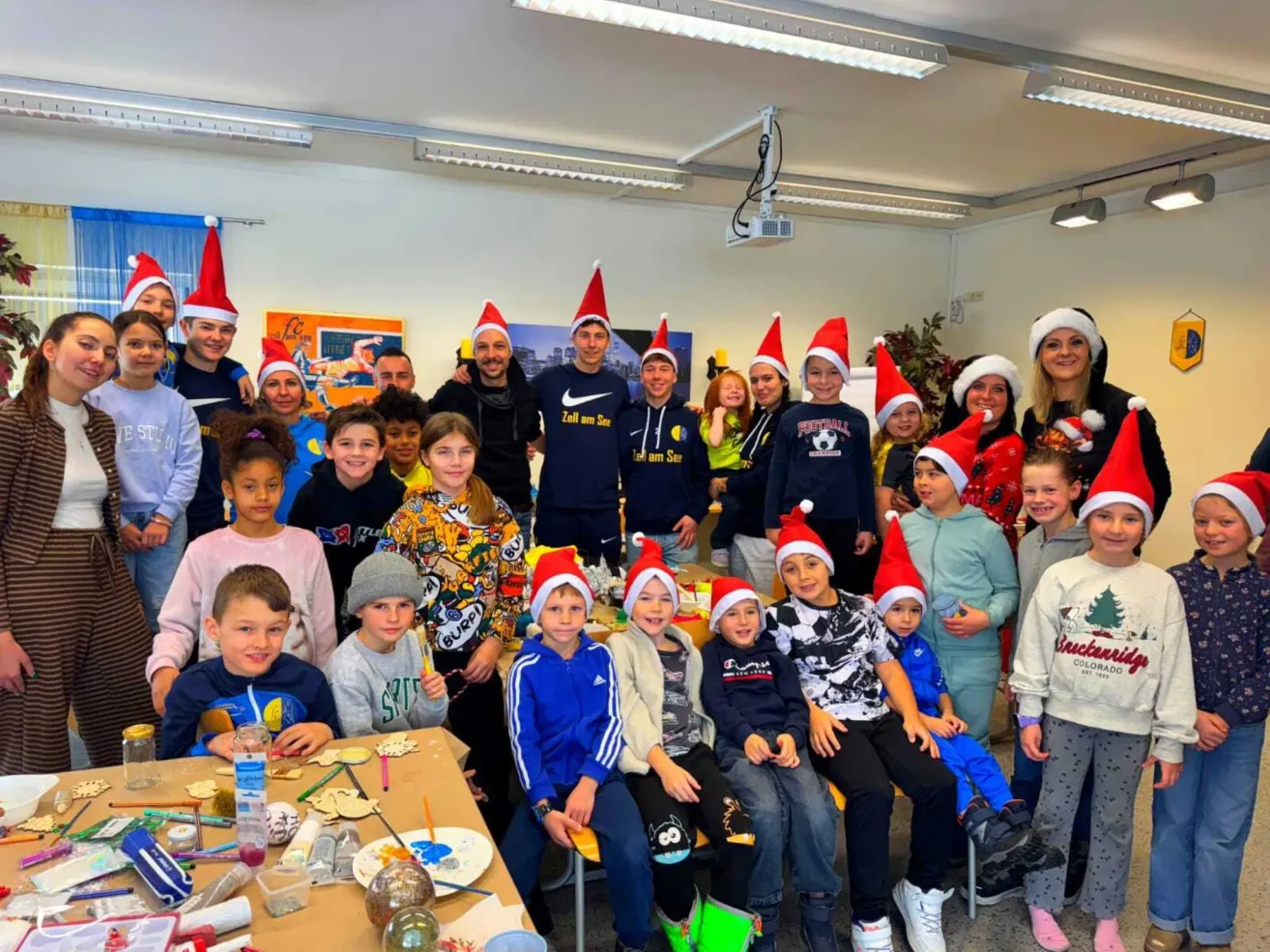Eine Gruppe von Kindern und Erwachsenen mit Weihnachtsmützen posiert für ein Foto in einem Raum mit Tischen und Bastelarbeiten.