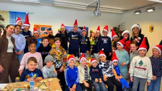 Eine Gruppe von Kindern und Erwachsenen mit Weihnachtsmützen posiert für ein Foto in einem Raum mit Tischen und Bastelarbeiten.