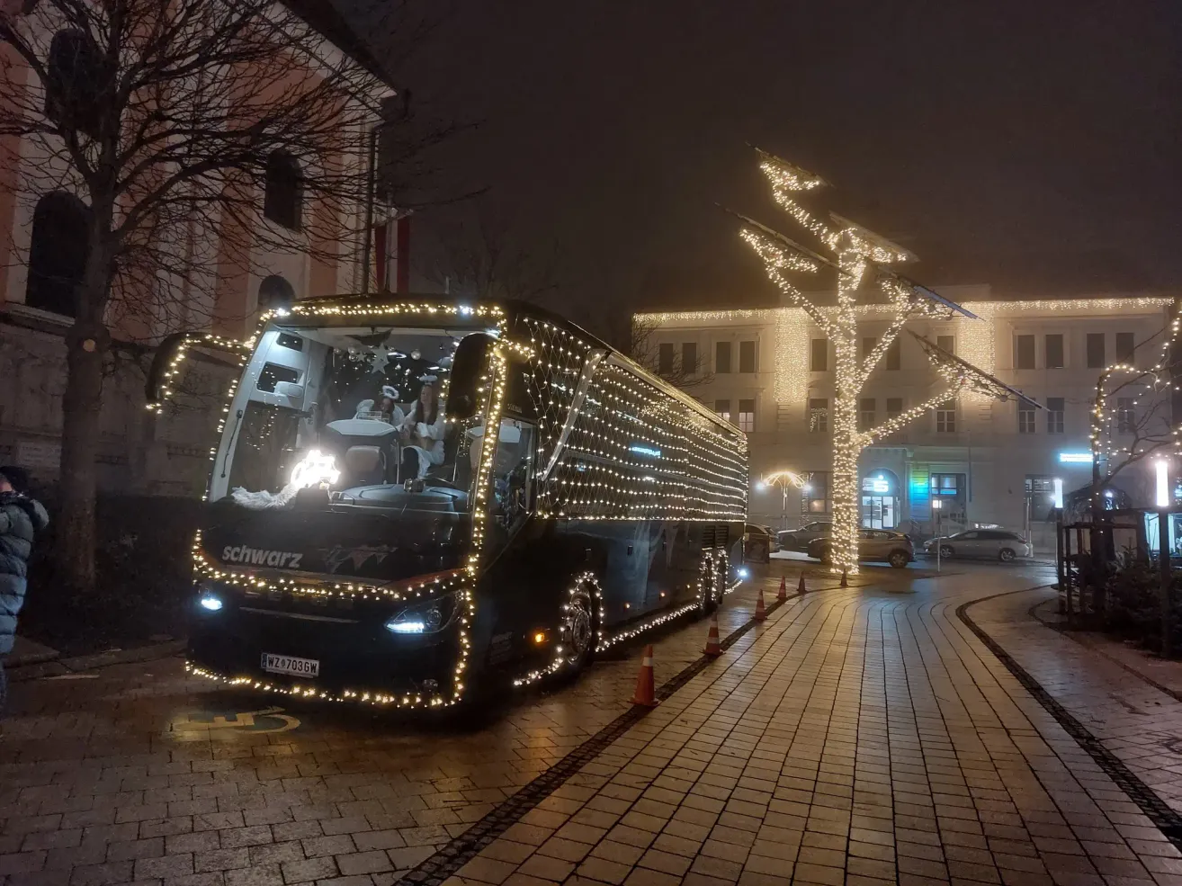 Ein mit Weihnachtslichtern geschmückter Bus ist nachts auf einer Straße geparkt. Der Bus hat das Wort schwarz auf seiner Seite. Auf dem Bürgersteig stehen zwei beleuchtete Bäume. Ein Gebäude befindet sich hinter den Bäumen. Vor dem Gebäude sind Autos geparkt.
