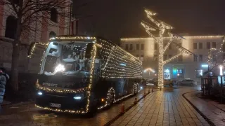 Ein mit Weihnachtslichtern geschmückter Bus ist nachts auf einer Straße geparkt. Der Bus hat das Wort schwarz auf seiner Seite. Auf dem Bürgersteig stehen zwei beleuchtete Bäume. Ein Gebäude befindet sich hinter den Bäumen. Vor dem Gebäude sind Autos geparkt.