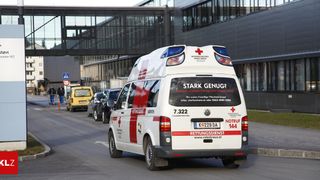 Ein weißer Krankenwagen mit roten Kreuzen fährt die Straße hinunter, im Hintergrund ein Gebäude. Auf der Rückseite des Krankenwagens steht "Stark Genug?" und hat ein rotes Kreuz auf der Heckklappe.