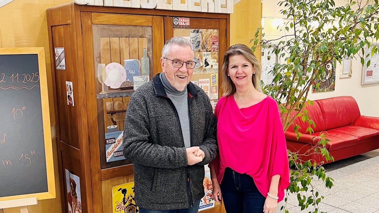 Ein älterer Mann und eine Frau stehen nebeneinander vor einer Vitrine mit verschiedenen Gegenständen. Der Mann trägt eine Brille und eine Jacke, während die Frau ein rosafarbenes Oberteil trägt. Hinter ihnen steht eine Pflanze.