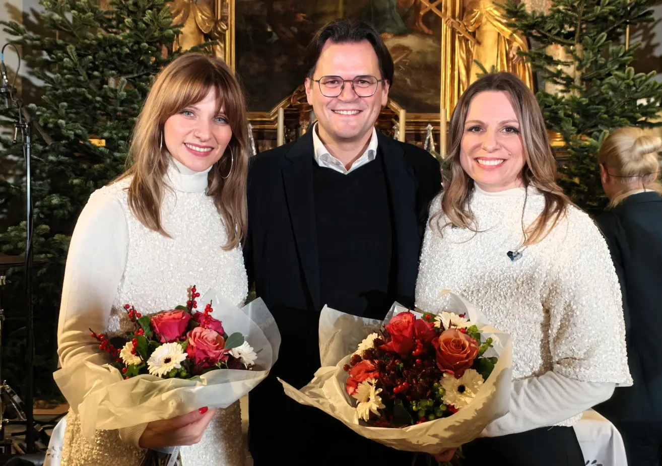 Drei Menschen lächeln für ein Foto. Ein Mann und zwei Frauen. Sie halten alle Blumen. Die Frau auf der linken Seite trägt ein weißes Kleid, die Frau auf der rechten Seite trägt ein weißes Oberteil. Der Mann trägt einen schwarzen Anzug.