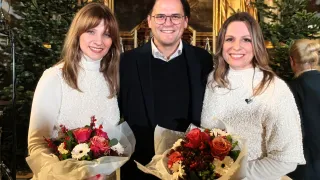 Drei Menschen lächeln für ein Foto. Ein Mann und zwei Frauen. Sie halten alle Blumen. Die Frau auf der linken Seite trägt ein weißes Kleid, die Frau auf der rechten Seite trägt ein weißes Oberteil. Der Mann trägt einen schwarzen Anzug.