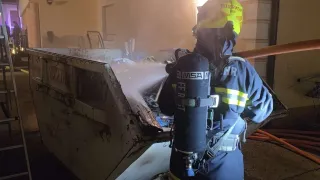 Ein Feuerwehrmann mit einem Helm, der mit 'Feuerwehr' beschriftet ist, und einem Feuerlöscher steht neben einem Müllcontainer und spritzt Wasser darauf.