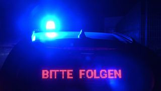 Das beleuchtete Schild auf einem Auto zeigt 'BITTE FOLGEN' mit einer blauen blinkenden Leuchte im Hintergrund.
