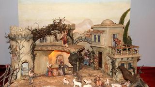 Eine Miniaturkrippe mit Figuren, Tieren und Gebäuden in einer ländlichen Landschaft. Figuren umfassen Jesus, Maria und Josef, umgeben von Hirten und Tieren.