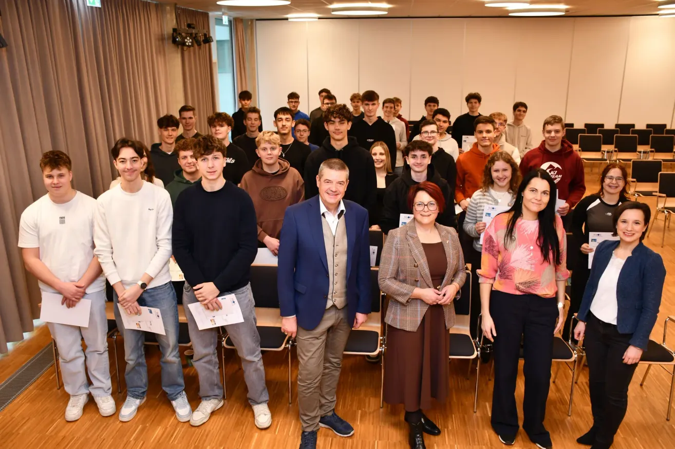 Eine Gruppe von Schülern steht in einem Raum mit einem Mann in einem blauen Anzug und einer Frau mit Brille. Sie alle haben Urkunden und lächeln. Stühle sind hinter ihnen aufgestellt, und der Raum ist gut beleuchtet.