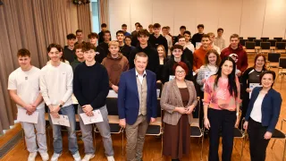 Eine Gruppe von Schülern steht in einem Raum mit einem Mann in einem blauen Anzug und einer Frau mit Brille. Sie alle haben Urkunden und lächeln. Stühle sind hinter ihnen aufgestellt, und der Raum ist gut beleuchtet.