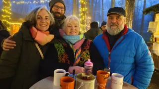 Vier Menschen lächeln für ein Foto in einem beleuchteten Waldgebiet. Ein Tisch mit Tassen und einem Wasserkocher ist im Vordergrund.