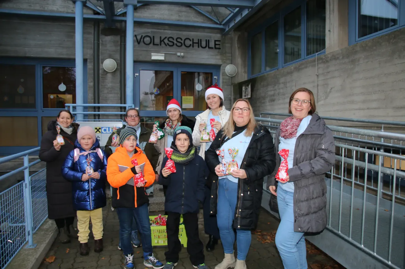 Eine Gruppe von Menschen, Erwachsene und Kinder, steht vor einem Gebäude mit dem Namen Volksschule. Sie tragen alle Winterkleidung und halten Geschenktüten. Einige tragen Weihnachtsmützen und Brillen. Die Frau in der Mitte hält eine Zuckerstange.