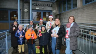 Eine Gruppe von Menschen, Erwachsene und Kinder, steht vor einem Gebäude mit dem Namen Volksschule. Sie tragen alle Winterkleidung und halten Geschenktüten. Einige tragen Weihnachtsmützen und Brillen. Die Frau in der Mitte hält eine Zuckerstange.