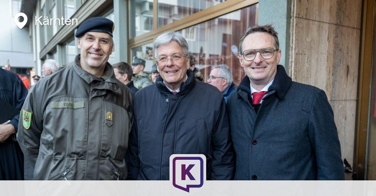 Drei Männer stehen lächelnd für ein Foto im Freien. Einer trägt eine Militärmütze, ein anderer ein rotes Halstuch. Ein Gebäude mit Glasfenstern und Soldaten im Hintergrund.