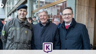 Drei Männer stehen lächelnd für ein Foto im Freien. Einer trägt eine Militärmütze, ein anderer ein rotes Halstuch. Ein Gebäude mit Glasfenstern und Soldaten im Hintergrund.