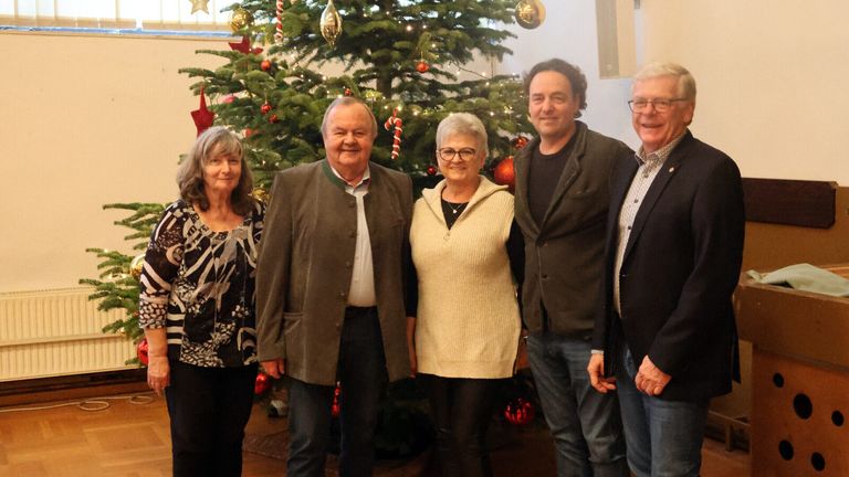 Fünf Personen, möglicherweise eine Familie, stehen vor einem geschmückten Weihnachtsbaum. Zwei Männer und drei Frauen, alle lächelnd, scheinen für ein Foto zu posieren.
