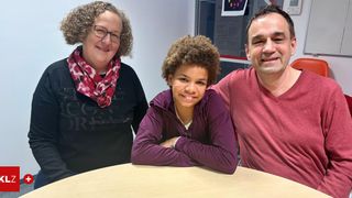 Drei Menschen, zwei Erwachsene und ein Kind, lächeln und posieren für ein Foto. Die Erwachsenen stehen hinter einem Tisch, und das Kind sitzt darauf.