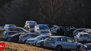 Eine Reihe von Autos und Rettungsfahrzeugen sind auf einem Parkplatz geparkt, umgeben von laublosen Bäumen. Eine Person in einem hellen gelben Vest steht neben einem weißen Van.