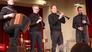 Drei Musiker treten auf der Bühne auf. Einer spielt Klarinette, der andere Akkordeon und der dritte ebenfalls Klarinette. Dahinter befindet sich ein roter Vorhang und Holzstühle.