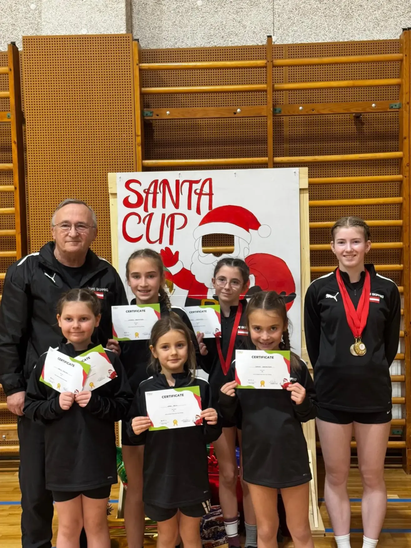 Eine Gruppe von Mädchen und zwei Erwachsene stehen vor einem Santa Cup-Banner. Sie halten Urkunden und Medaillen und lächeln für ein Foto.