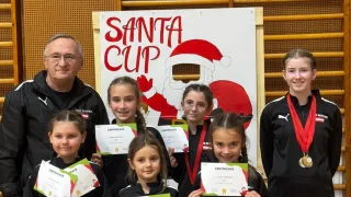 Eine Gruppe von Mädchen und zwei Erwachsene stehen vor einem Santa Cup-Banner. Sie halten Urkunden und Medaillen und lächeln für ein Foto.