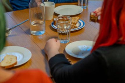 Zwei Menschen mit roter und grüner Haarfarbe sitzen am Holztisch und essen. Teller, Gläser und Tassen stehen auf dem Tisch.