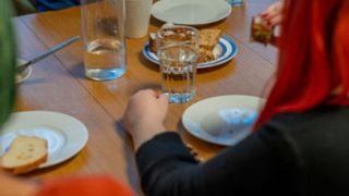 Zwei Menschen mit roter und grüner Haarfarbe sitzen am Holztisch und essen. Teller, Gläser und Tassen stehen auf dem Tisch.