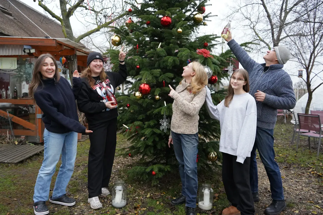 Eine Gruppe von Menschen steht um einen geschmückten Weihnachtsbaum herum, hält Ornamente und Kerzen. Ein Mann hält eine Flasche, während ein anderer zum Baum aufblickt.