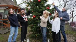 Eine Gruppe von Menschen steht um einen geschmückten Weihnachtsbaum herum, hält Ornamente und Kerzen. Ein Mann hält eine Flasche, während ein anderer zum Baum aufblickt.