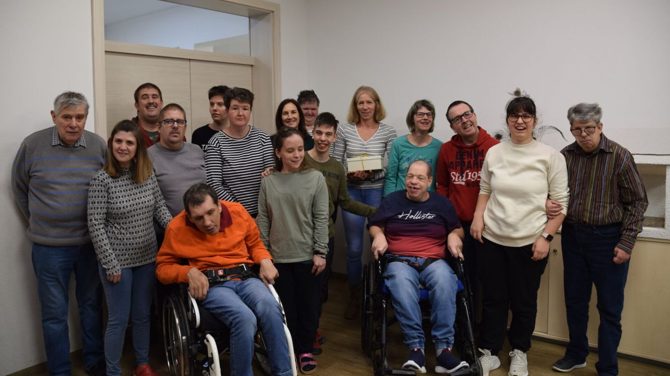 Eine Gruppe von Personen, darunter einige in Rollstühlen, lächelt und posiert für ein Foto. Eine Person in der Mitte hält eine Geschenkbox. Eine andere Person trägt eine rote Kapuzenjacke mit Text. Eine weiße Wand ist hinter ihnen.