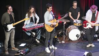Drei Musiker treten auf einer Bühne auf. Ein Mann spielt eine Gitarre und singt in ein Mikrofon. Eine Frau sitzt auf einem Hocker und spielt eine Tastatur. Ein anderer Mann spielt ein Schlagzeug.