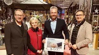 Vier Personen stehen in einem dekorierten Raum und halten ein Schild, das '#STRONG' und '3.000€' zeigt. Der Mann in der Mitte trägt einen schwarzen Anzug und hält das Schild, lächelt. Die Frau neben ihm trägt eine rote Jacke. Dahinter hängen Ornamente und Lichter.