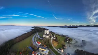 Luftaufnahme eines friedlichen Dorfes in einem nebligen Tal mit einer majestätischen Burg in seiner Mitte, umgeben von einer üppigen grünen Landschaft und einem klaren blauen Himmel.