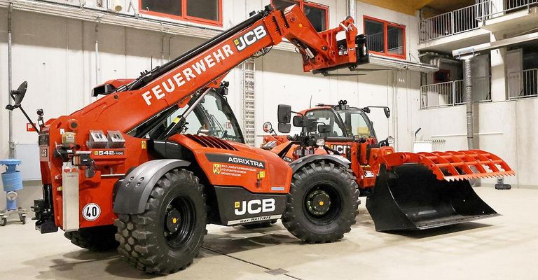Zwei orange JCB-Landwirtschaftstraktoren sind in einem Lagerhaus geparkt. Beide haben große Reifen und angehobene Arme. Einer hat einen Pflug befestigt.