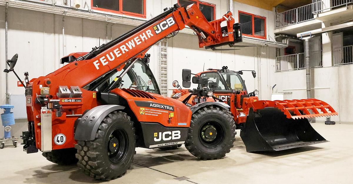 Zwei orange JCB-Landwirtschaftstraktoren sind in einem Lagerhaus geparkt. Beide haben große Reifen und angehobene Arme. Einer hat einen Pflug befestigt.