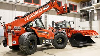 Zwei orange JCB-Landwirtschaftstraktoren sind in einem Lagerhaus geparkt. Beide haben große Reifen und angehobene Arme. Einer hat einen Pflug befestigt.