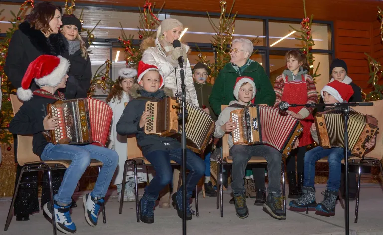 Eine Gruppe von Kindern und Erwachsenen spielt Akkordeons und singt vor einem Mikrofon mit Weihnachtsdekoration im Hintergrund.