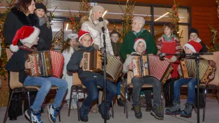 Eine Gruppe von Kindern und Erwachsenen spielt Akkordeons und singt vor einem Mikrofon mit Weihnachtsdekoration im Hintergrund.