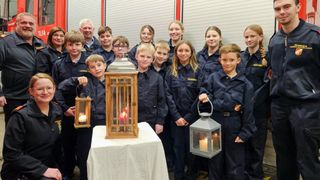 Eine Gruppe von Kindern in Marineuniformen, die Laternen mit brennenden Kerzen halten, stehen in einer Reihe. Dahinter steht ein Tisch mit einer größeren Laterne mit brennenden Kerzen.