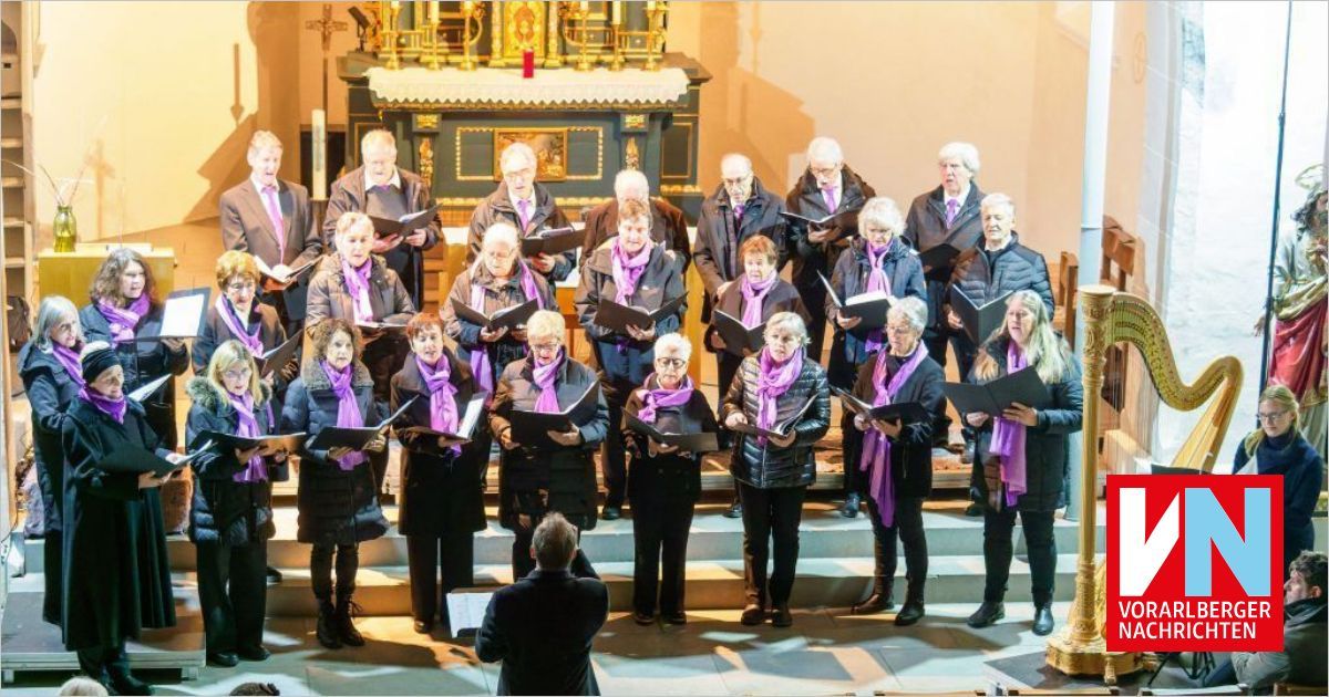 Ein Chor in lila Schals singt auf einer Bühne vor einem Altar. Der Dirigent steht vorne, und die Chormitglieder halten Notenblätter in den Händen.