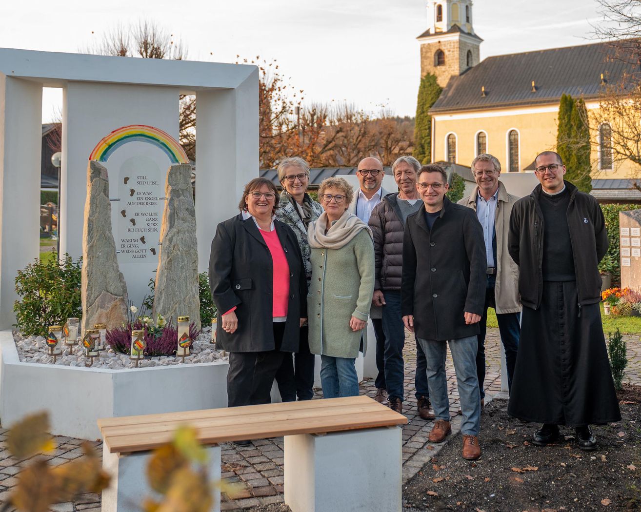 Eine Gruppe von Menschen steht vor einem Denkmal in der Nähe einer Kirche auf einem Friedhof. Sie lächeln und posieren für ein Foto. Das Denkmal hat ein Regenbogendesign und einige darauf geschriebene Texte.