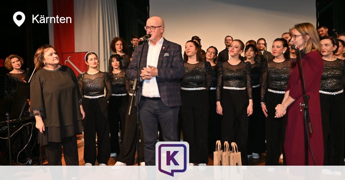 Ein Chor steht auf der Bühne mit einem Mann, der in ein Mikrofon spricht. Die Chormitglieder tragen schwarze Outfits mit silbernen Streifen.