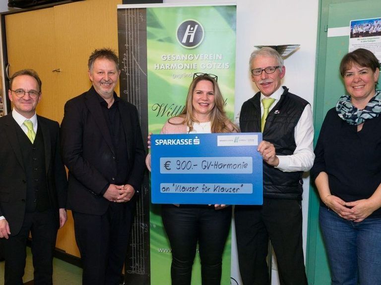 Fünf Personen stehen eng beieinander in einem Raum. Eine Frau hält einen blauen Scheck mit der Aufschrift Sparkasses und €900-GV-Harmonie. Sie lächelt in die Kamera. Hinter ihr steht ein Plakat, auf dem Gesangverein Harmonie Gotzis steht.