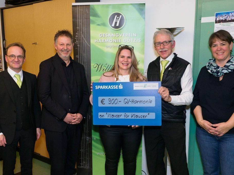 Fünf Personen stehen eng beieinander in einem Raum. Eine Frau hält einen blauen Scheck mit der Aufschrift Sparkasses und €900-GV-Harmonie. Sie lächelt in die Kamera. Hinter ihr steht ein Plakat, auf dem Gesangverein Harmonie Gotzis steht.